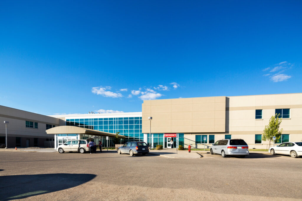 Exterior view of a modern medical facility with a prominent Emergency entrance and a covered driveway, demonstrating the accessible front-door parking solutions required at busy hospitals.